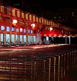 A row of ticket booths under a red and white striped canopy is lit with warm lights. The booths display digital screens and are arranged along metal railings. The scene is illuminated in a night setting with an empty path leading towards the booths.