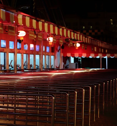 A row of ticket booths under a red and white striped canopy is lit with warm lights. The booths display digital screens and are arranged along metal railings. The scene is illuminated in a night setting with an empty path leading towards the booths.
