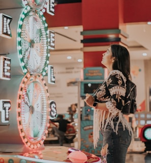 A woman is standing in an arcade looking at a large colorful game machine with bright lights. The game machine features large spinning wheels labeled with 'tickets' and has a vibrant design. The woman is casually dressed in a jacket with fringe details and is holding something in her hand while wearing a smartwatch.