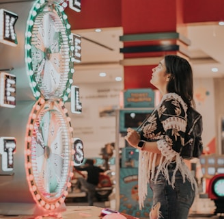 A woman is standing in an arcade looking at a large colorful game machine with bright lights. The game machine features large spinning wheels labeled with 'tickets' and has a vibrant design. The woman is casually dressed in a jacket with fringe details and is holding something in her hand while wearing a smartwatch.