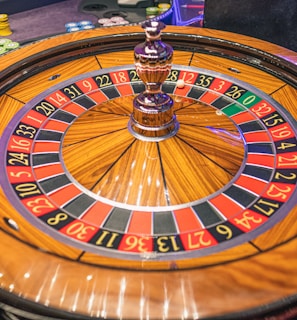 A roulette wheel with alternating red and black numbered pockets on a wooden platform. The wheel contains a small white ball resting in one of the pockets, indicating gameplay. Surrounding the wheel are stacks of colorful poker chips, suggesting a casino environment.