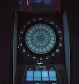 A digital dartboard is mounted within a dimly lit space, featuring a circular target labeled with numbered sections from 1 to 20. Below the target area, there are control panels with several buttons labeled P1 to P4, a start button, and additional controls. Above the dartboard, a screen displays a faint projection of an image, possibly of a game interface or other electronic display.
