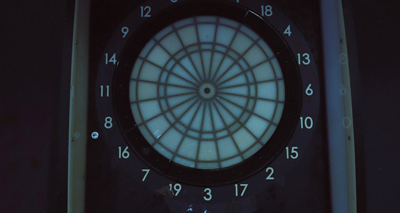 A digital dartboard is mounted within a dimly lit space, featuring a circular target labeled with numbered sections from 1 to 20. Below the target area, there are control panels with several buttons labeled P1 to P4, a start button, and additional controls. Above the dartboard, a screen displays a faint projection of an image, possibly of a game interface or other electronic display.